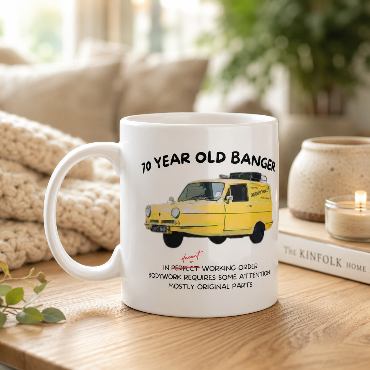 A white ceramic mug with a printed illustration of a yellow vintage car and the text '40 YEAR OLD BANGER' and a quote, displayed on a wooden table in a cozy living room setting.