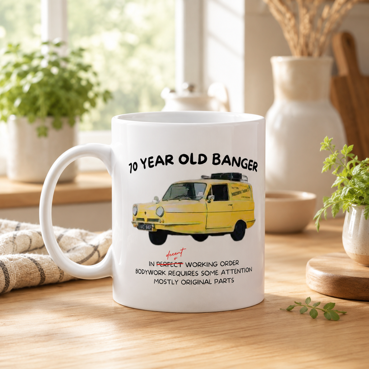 A white ceramic coffee mug with a printed illustration of a yellow vintage van and the text '50 YEAR OLD BANGER' and 'IN PERFECT WORKING ORDER BODYWORK REQUIRES SOME ATTENTION MOSTLY ORIGINAL PARTS', shown on a wooden table in a kitchen setting.