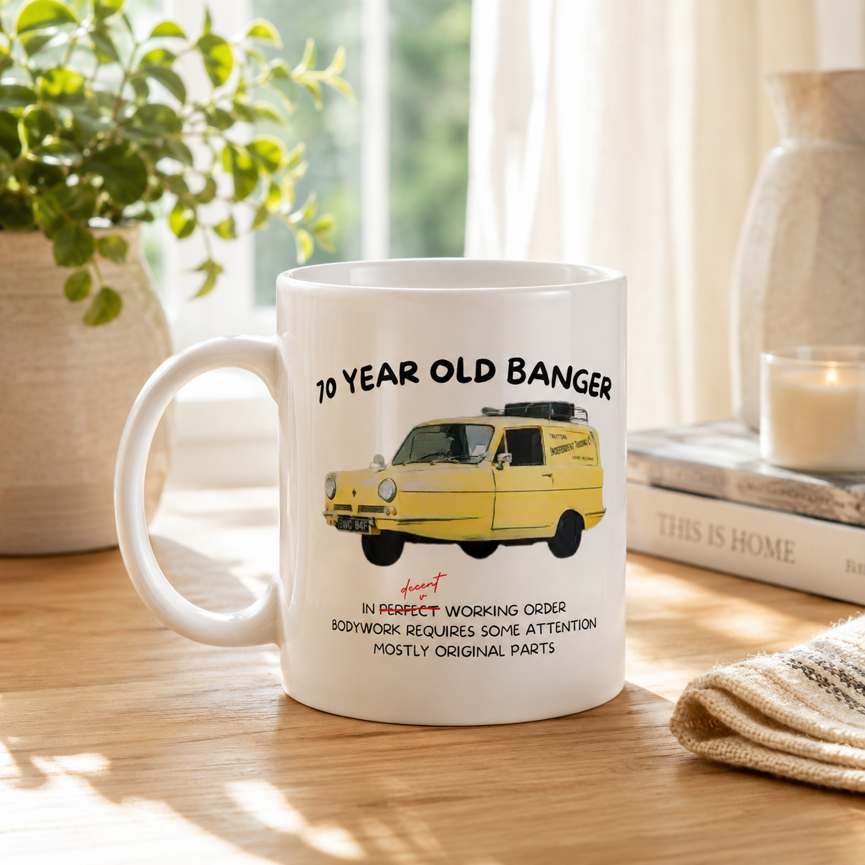 A white ceramic mug with a yellow vintage van graphic and the text '60 YEAR OLD BANGER' sits on a wooden table in a softly lit room with books, a plant, and a candle in the background.