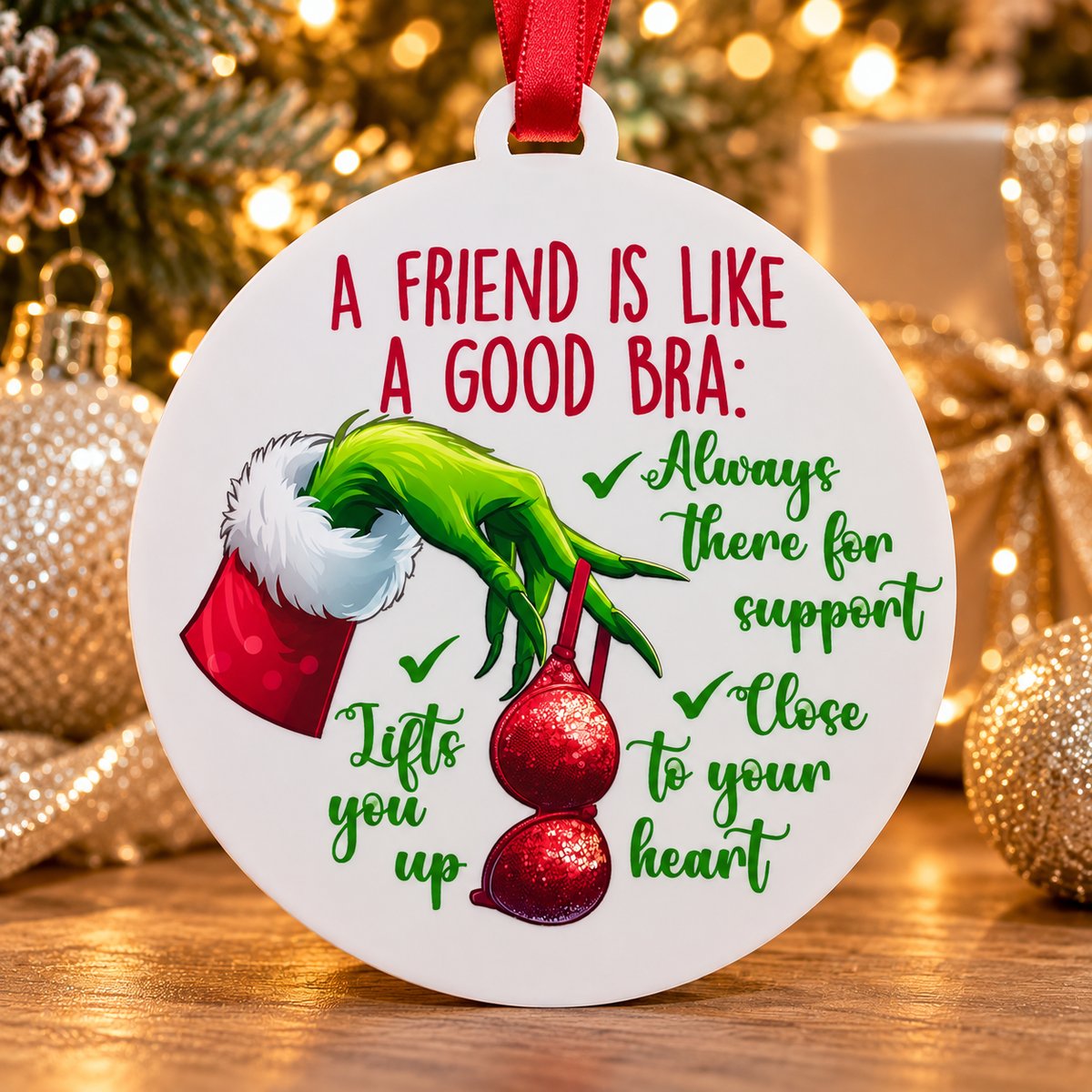 A round white Christmas ornament with a red hanging ribbon, featuring a green hand reaching out of a red Santa hat and red ornaments, with the text 'A FRIEND IS LIKE A GOOD BRA: Always there for support, Lifts you up, Close to your heart' on a festive blurred holiday background.