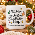 A white ceramic mug with a humorous Christmas message, displayed on a rustic wood slice surrounded by festive holiday decorations including red ornaments, pine branches, and warm string lights.