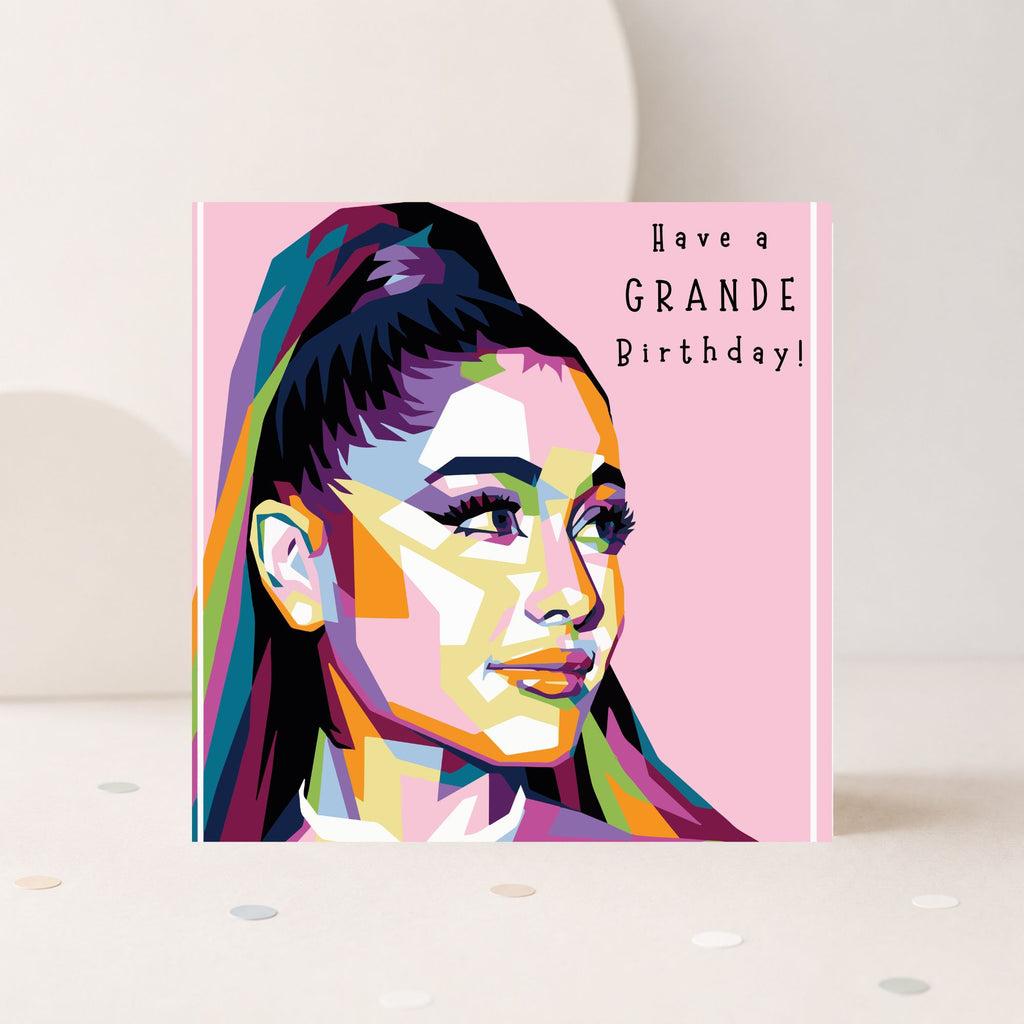 A square birthday card featuring a colorful pop-art portrait of Ariana Grande on a pink background, with the text 'Have a GRANDE Birthday!' printed on the right side. The card is standing upright on a light surface with confetti scattered around it.