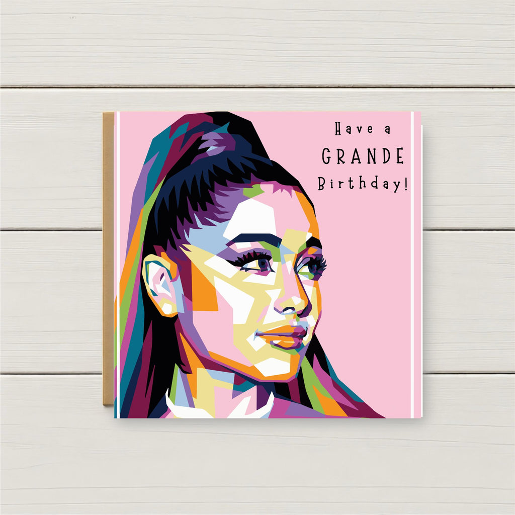 A square birthday card featuring a colorful pop-art portrait of a woman on a pink background with the text 'Have a GRANDE Birthday!' in black, photographed standing against a light wood plank surface.