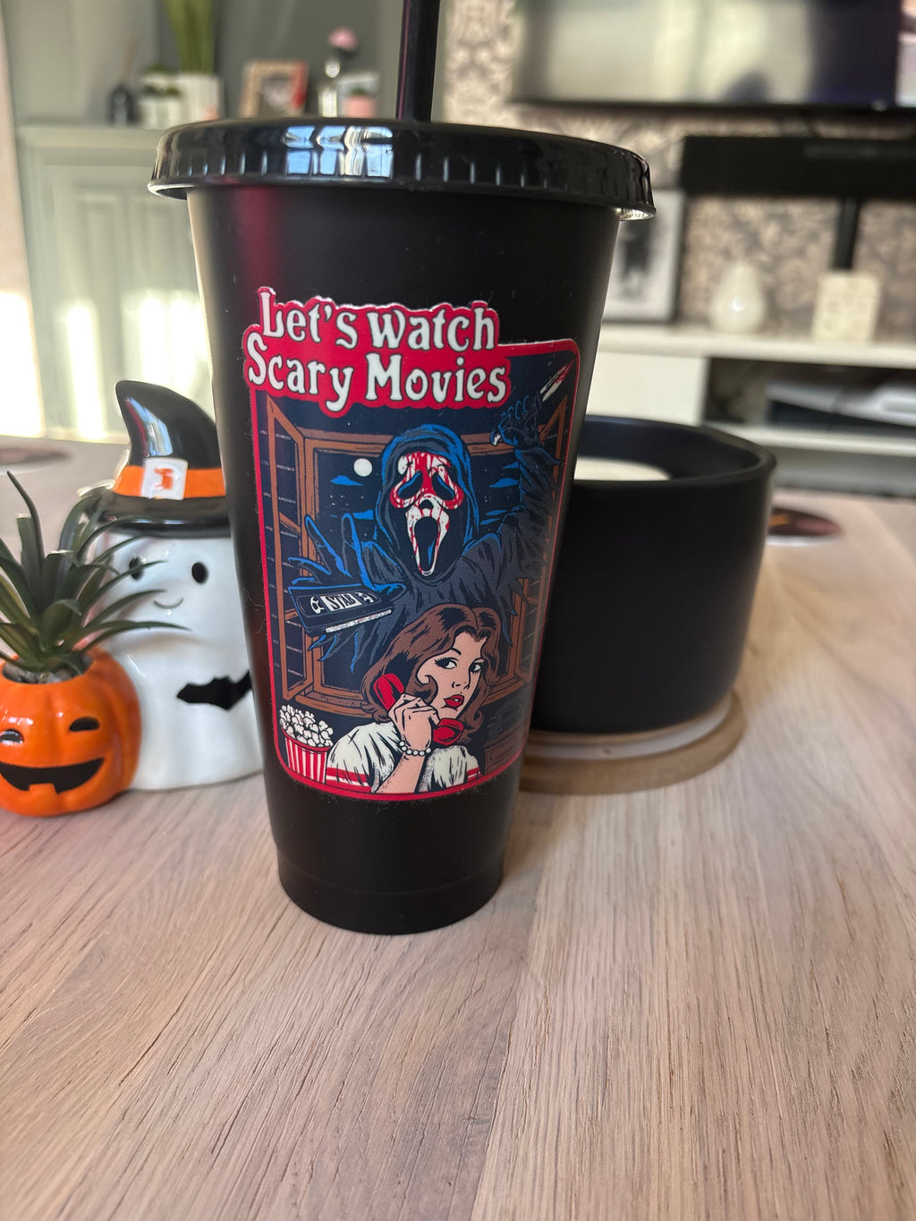 A black plastic travel tumbler with a lid and straw, featuring a colorful graphic of the Ghostface from the movie 'Let's Watch Scary Movies' and a woman on the phone. The tumbler is sitting on a light wood table in a home setting.