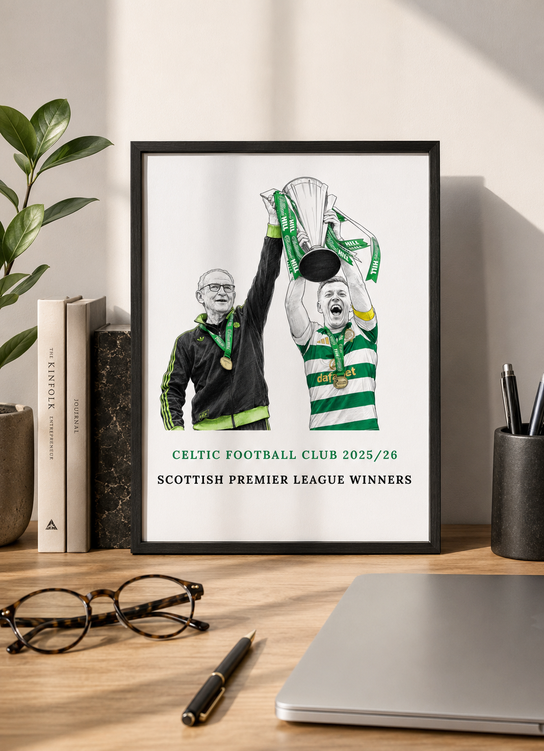 Framed poster of Celtic Football Club celebrating a Scottish Premier League win on a desk with books and a laptop.