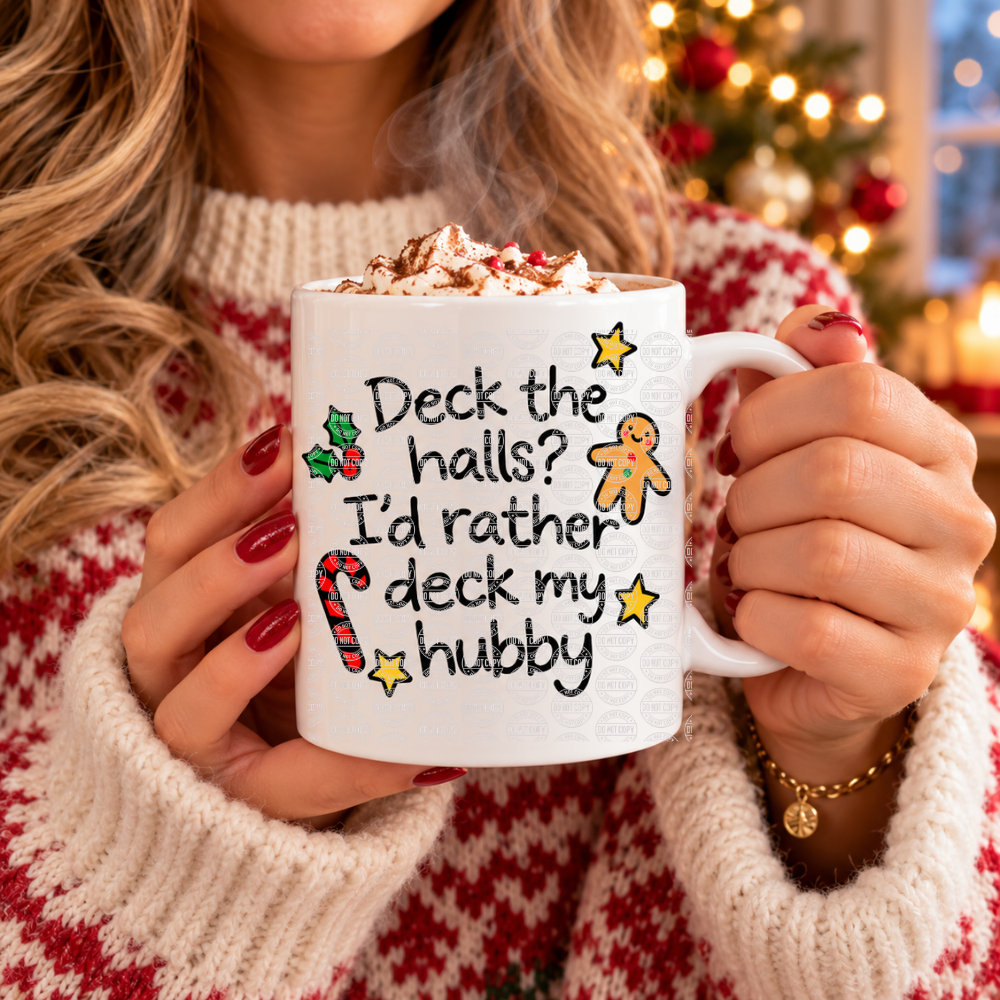 A person in a red and white knit sweater holds a white ceramic mug topped with whipped cream and cocoa. The mug has a Christmas-themed printed message reading 'Deck the halls? I'd rather deck my hubby' with candy cane, holly, gingerbread man, and star illustrations.