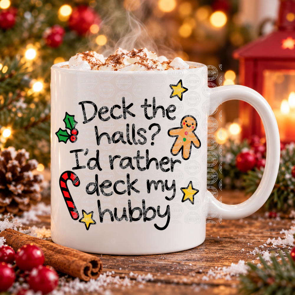 A white ceramic mug filled with a hot cocoa drink topped with whipped cream and cocoa powder, placed on a rustic wooden surface with Christmas decorations in the background. The mug is printed with the phrase 'Deck the halls? I'd rather deck my hubby' in black script, decorated with a gingerbread man, candy cane, holly, and yellow stars.