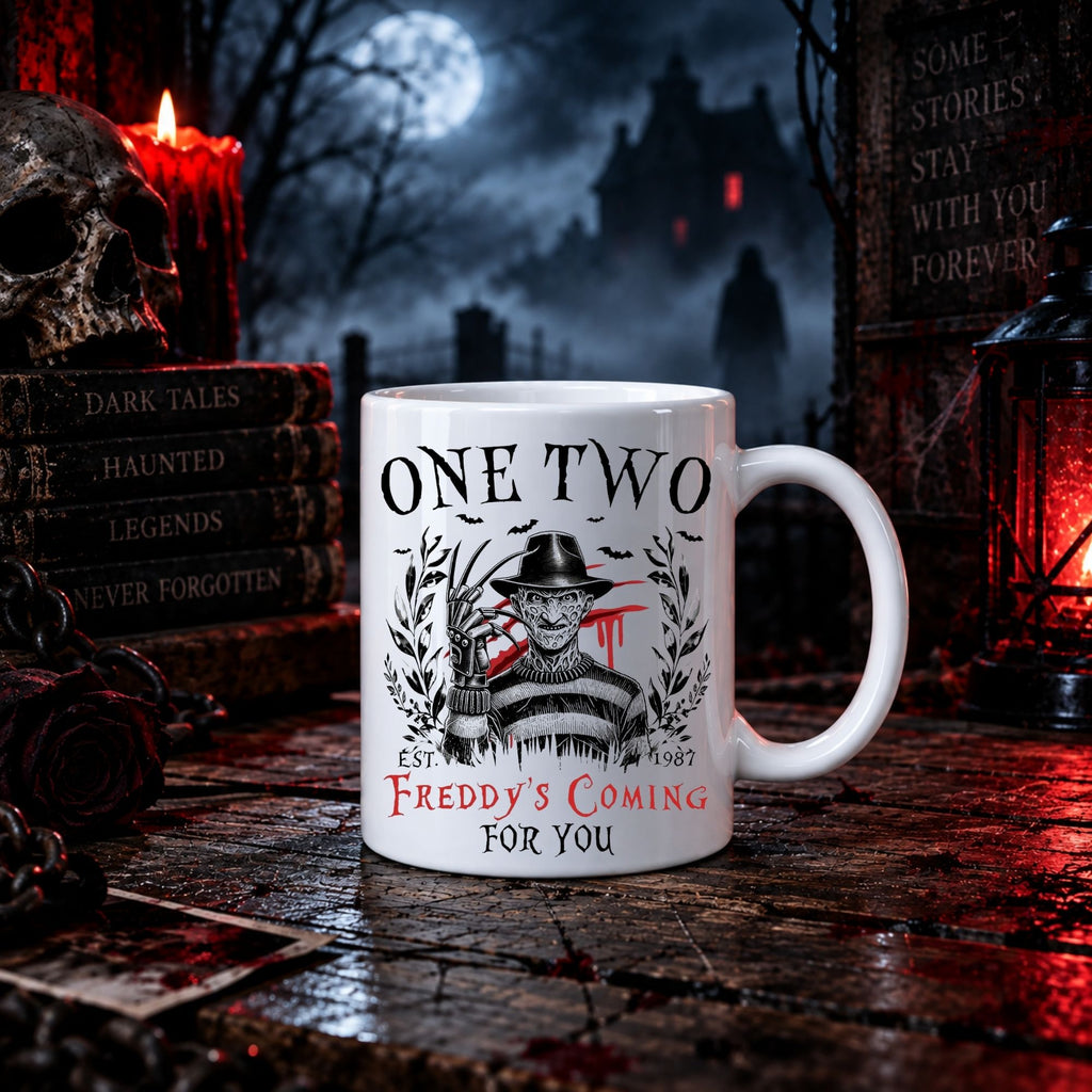 A white ceramic mug with a Freddy Krueger-themed graphic reading 'ONE TWO' and 'FREDDY'S COMING FOR YOU', styled on a dark wooden surface with Halloween decor including candles, a skull, books, and a lantern in the background.