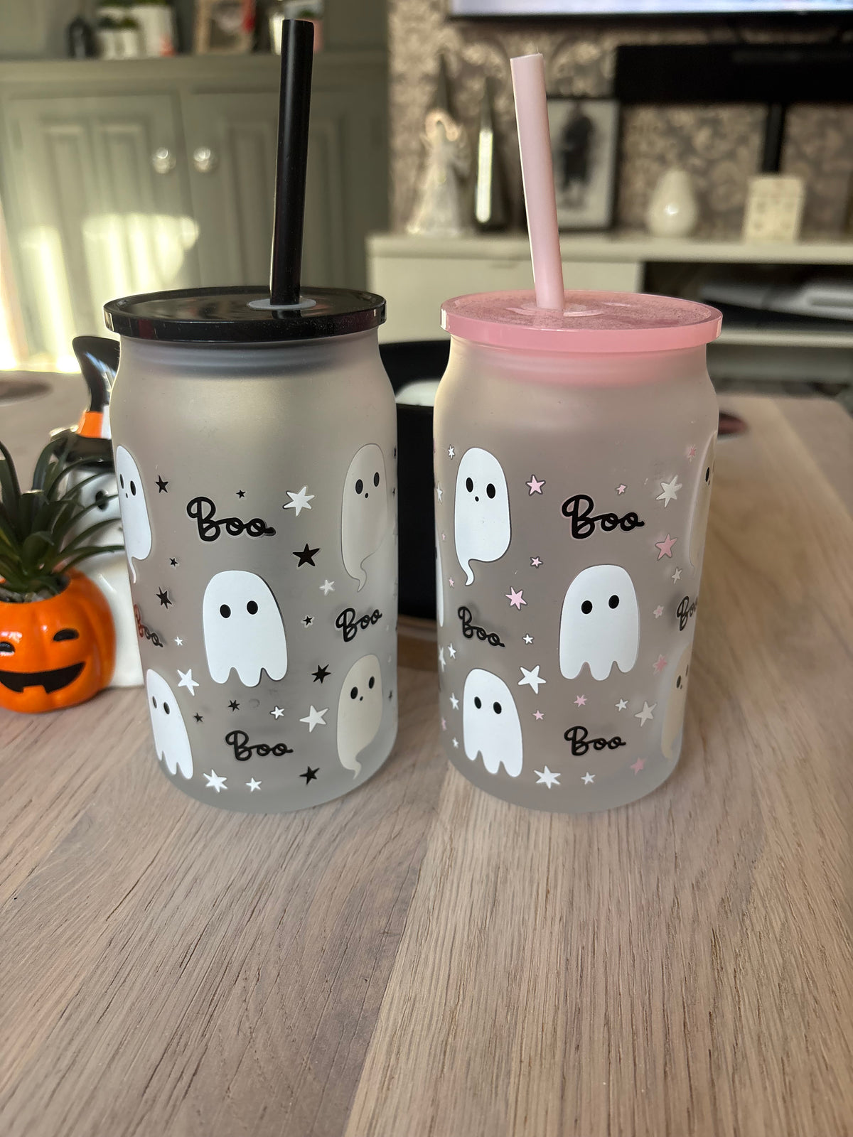 Two frosted Halloween-themed tumblers on a wooden table. The left tumbler has a black lid and straw, decorated with white ghosts, black stars, and the word 'Boo'.