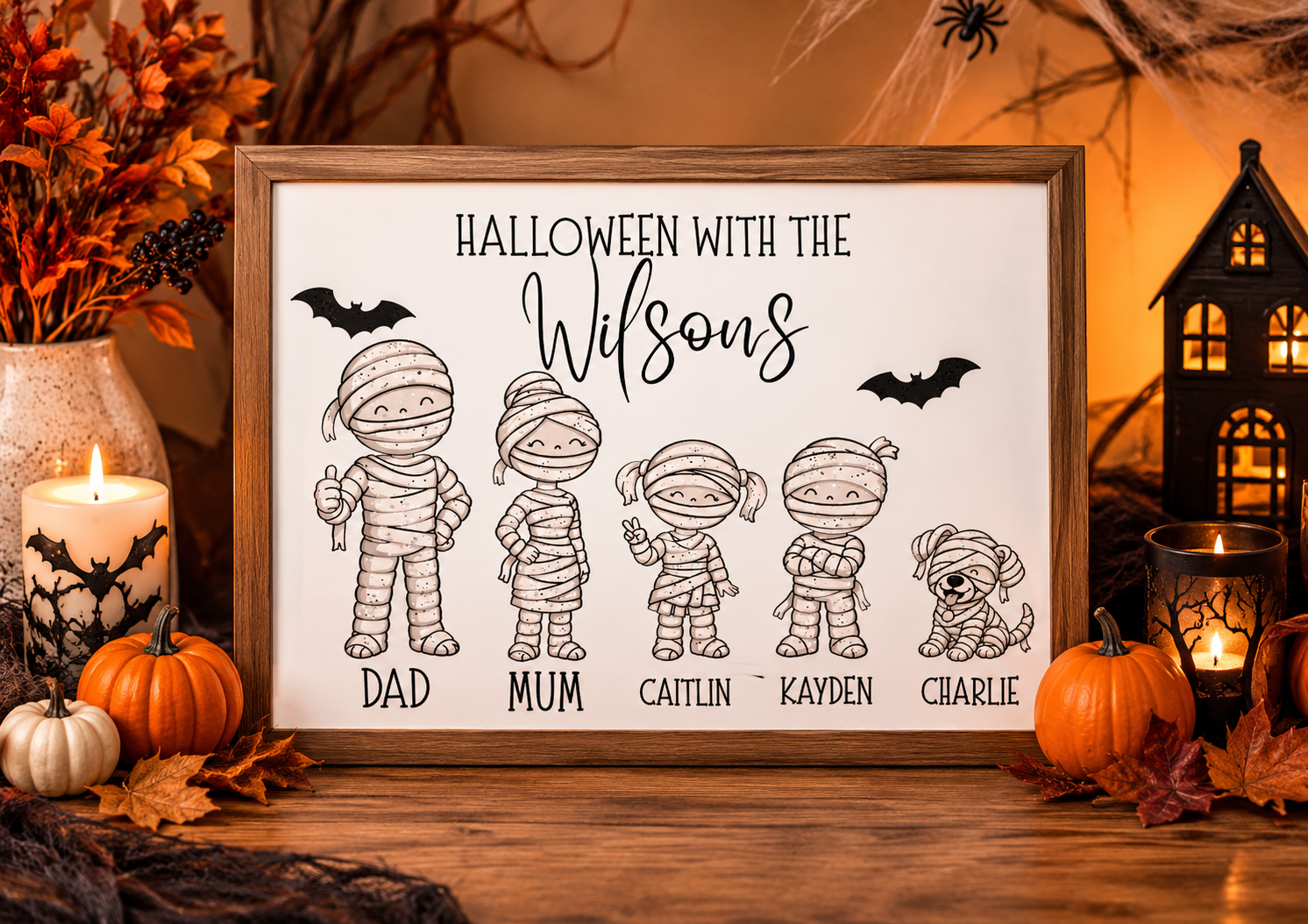 A framed Halloween sign with the text 'HALLOWEEN WITH THE Wilson's' and cartoon mummy figures labeled Dad, Mum, Caitlin, Kayden, and Charlie, displayed on a wooden surface with candles, pumpkins, autumn leaves, and a lantern in a warm fall setting.