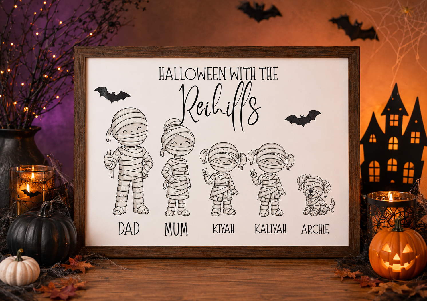 A framed Halloween-themed sign displayed on a wooden surface, surrounded by seasonal decor including pumpkins, candles, spiderwebs, and a haunted house silhouette. The sign reads “HALLOWEEN WITH THE Rehill’s” above black line drawings of five mummy-style figures labeled Dad, Mum, Kiyah, Kaliya, and Archie.