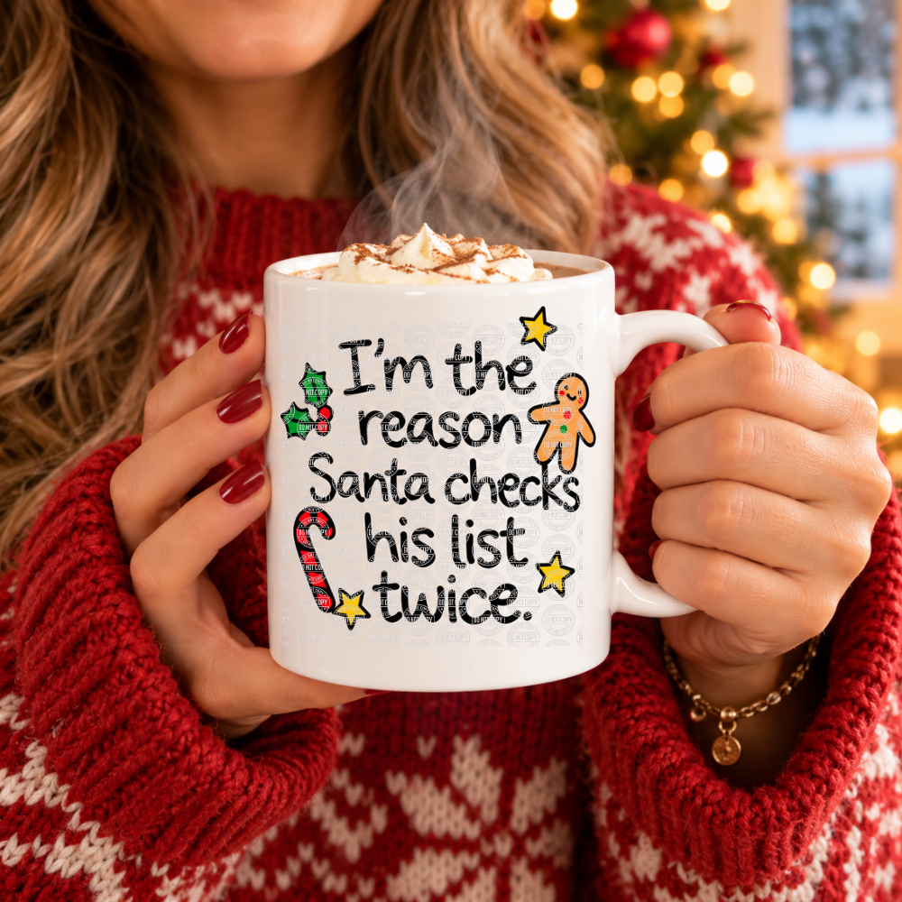 A person in a red knit sweater holds a white ceramic mug topped with whipped cream and cocoa, printed with the phrase “I’m the reason Santa checks his list twice.” with holiday illustrations.