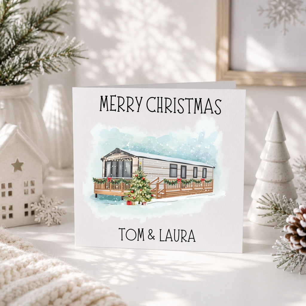 A personalized Christmas greeting card featuring a watercolor illustration of a holiday caravan with a decorated tree, displayed on a tabletop with festive props like a small house, pine branches, and a textured blanket.