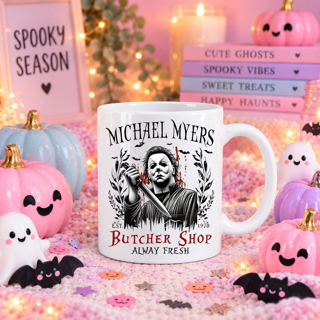 A white ceramic mug with a Halloween-themed Michael Myers butcher shop graphic, displayed in a cozy, pink-toned autumn setting with decorative pumpkins, candles, and themed signs.