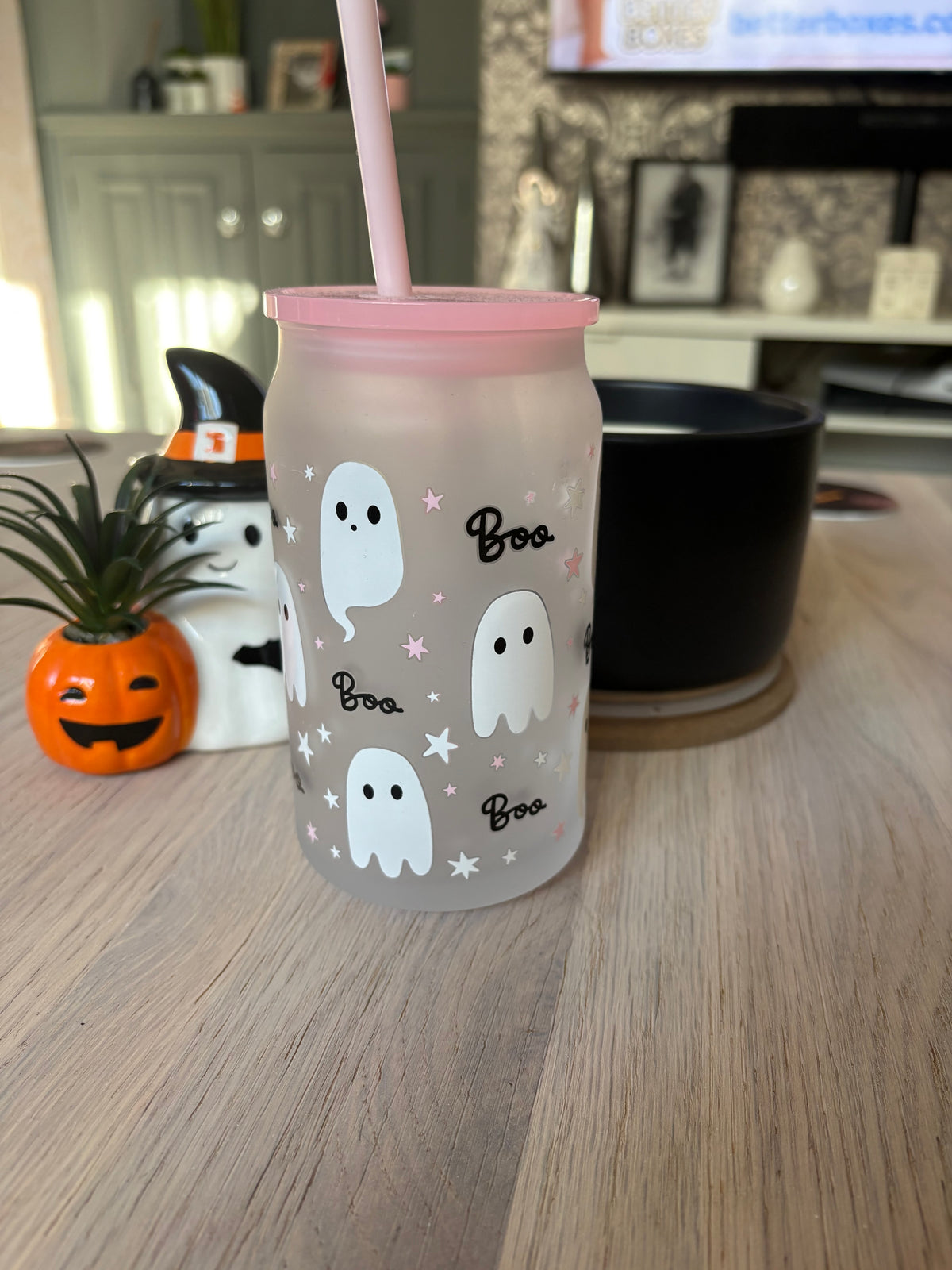 A frosted glass tumbler with a pink lid and straw, decorated with white cartoon ghosts, black 'Boo' text, and small pink stars, sitting on a light wood table in a living room setting.