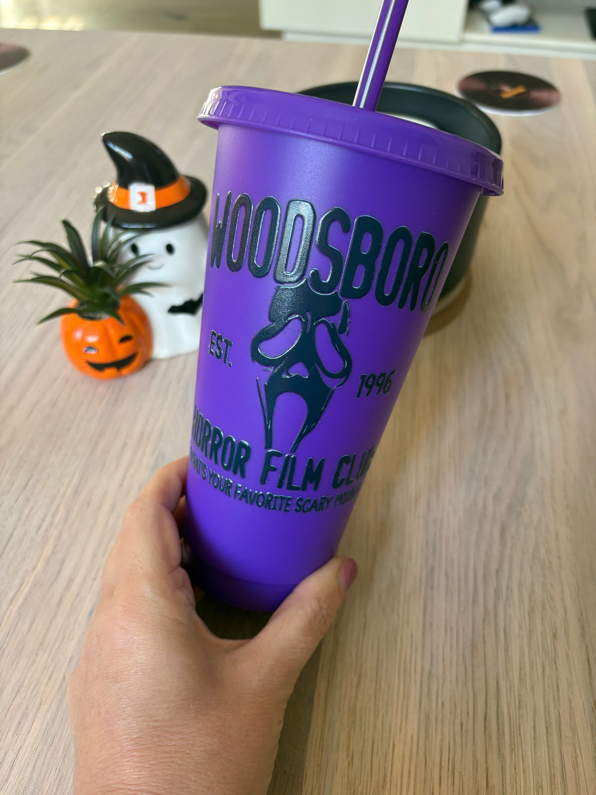A hand holds a purple Halloween-themed tumbler cup with a matching purple straw. The cup features a black Ghostface silhouette and the text 'HOODSBORO HORROR FILM CLUB EST.