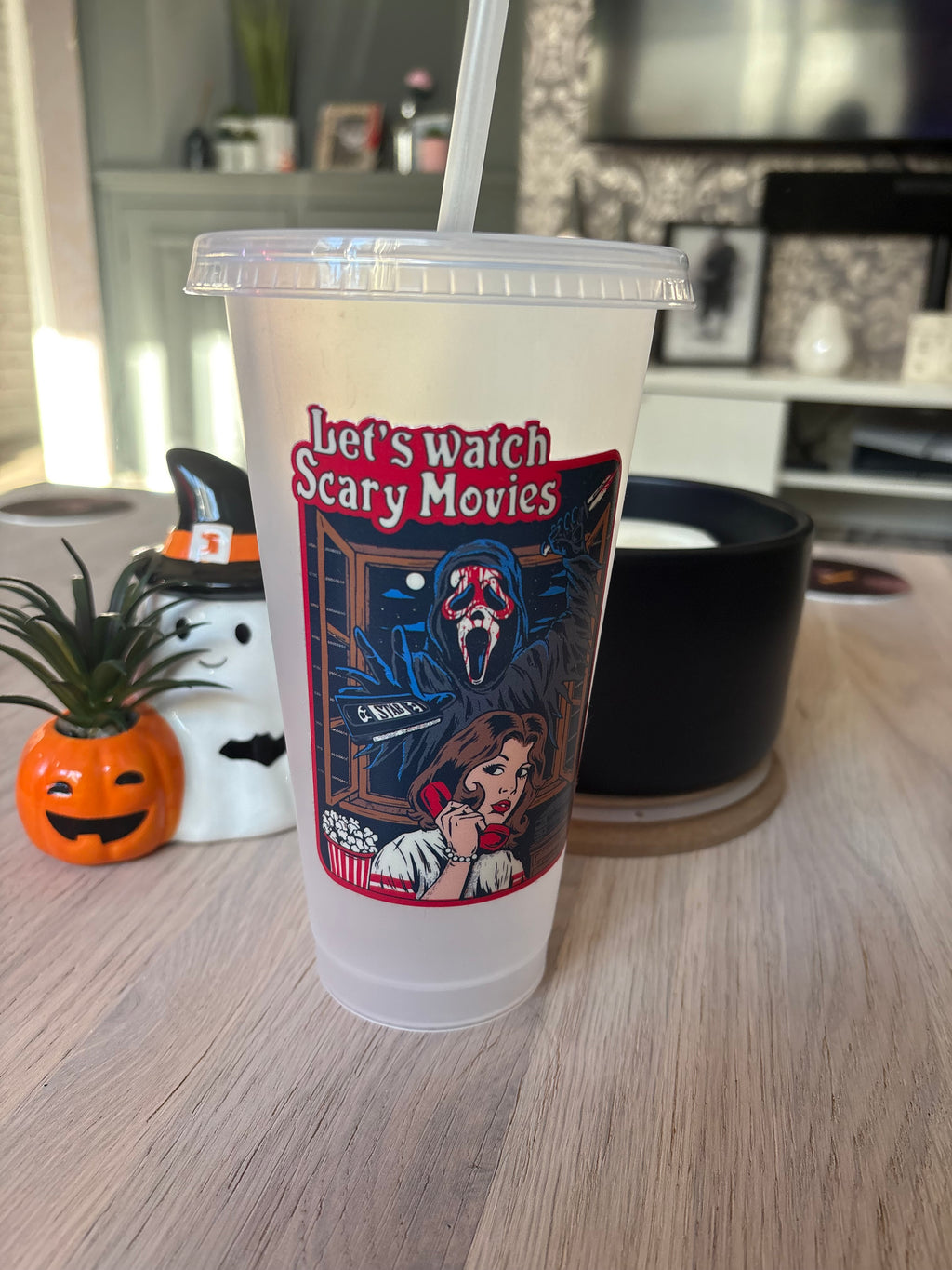 A clear plastic travel tumbler with a lid and straw, featuring a Halloween-themed graphic that reads 'Let's watch Scary Movies' with a ghost and a woman, set on a light wood table in a home environment.