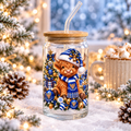 A clear glass jar tumbler with a cork lid and curved glass straw, decorated with a festive illustration of a Highland cow in a blue and white Santa hat and scarf, surrounded by ornaments and a Christmas tree. The tumbler is set in a snowy, holiday-themed scene with pine branches, lights, and a mug in the background.