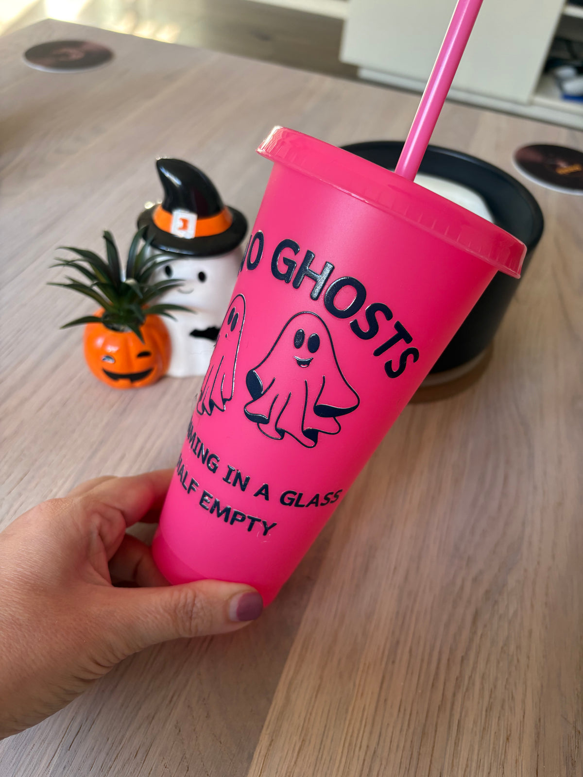 A hand holds a bright pink plastic tumbler with a matching pink straw. The cup has black text reading 'NO GHOSTS' and 'EMPTY IN A GLASS HALF EMPTY' with a ghost graphic.