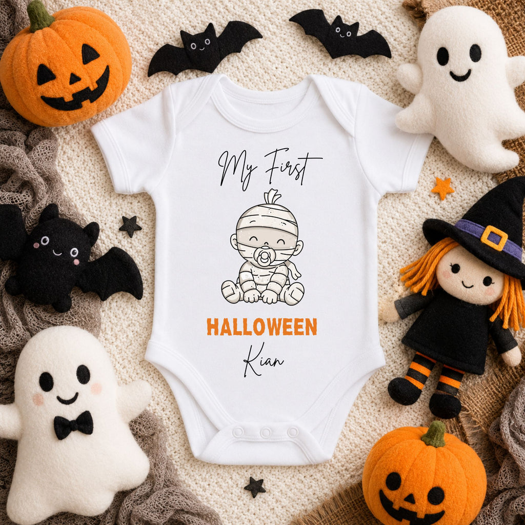 A white baby bodysuit with a Halloween-themed graphic print reading 'My First HALLOWEEN Kim' and showing a mummy sitting down, styled in a flat lay with plush ghosts, pumpkins, bats, and a witch doll around it.
