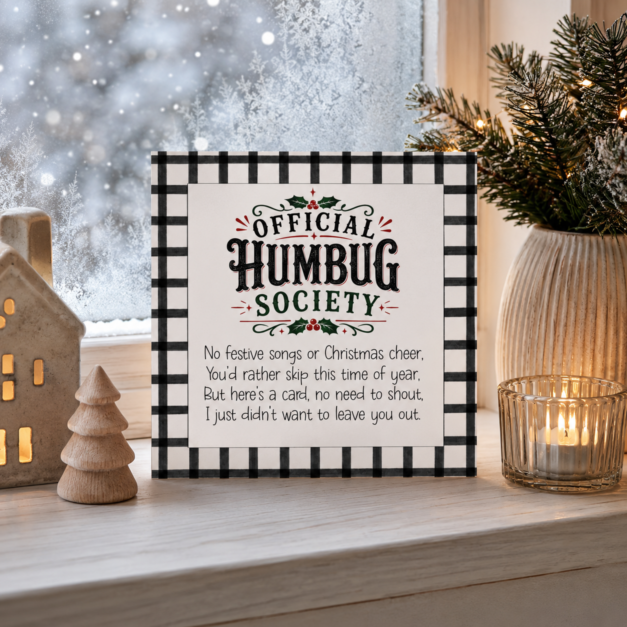 A square Christmas card with a black-and-white gingham border and the text 'OFFICIAL HUMBUG SOCIETY' and a humorous poem, styled on a windowsill with holiday decor including a lit candle, wooden tree, and snowy window view.