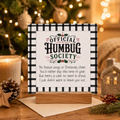 A square printed card with a black-and-white gingham border and the text 'OFFICIAL HUMBUG SOCIETY' and a short poem, displayed on a small wooden stand in a cozy holiday setting with a blurred Christmas tree and warm lights in the background.