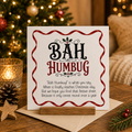 Bah Humbug cheeky Christmas card with holly design, standing on wooden card holder with festive background