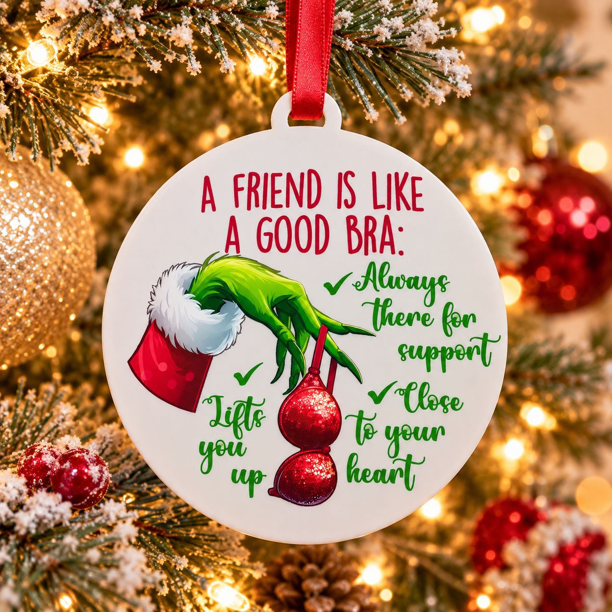 A round white Christmas ornament hanging from a red ribbon, featuring a green Grinch-like hand holding red holiday candies and the text 'A FRIEND IS LIKE A GOOD BRA: Always There for support, Lifts you up, Close to your heart.' The ornament is photographed in front of a lit Christmas tree with warm bokeh lights.