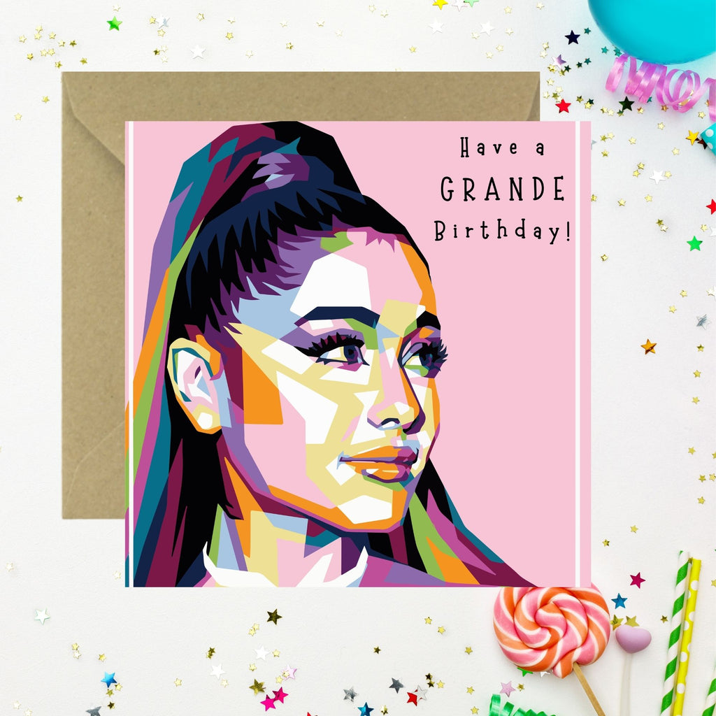 A square birthday card with a colorful pop-art portrait of Ariana Grande on a pink background, reading 'Have a GRANDE Birthday!' in black text. The card is shown with a brown envelope behind it on a white surface decorated with confetti, star stickers, a lollipop, and balloons.