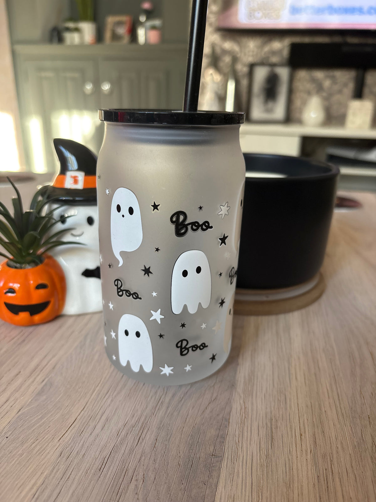 A frosted glass can-style tumbler with a black lid and straw, decorated with white ghosts, black stars, and the word 'Boo' in script, sitting on a wooden table in a home setting.