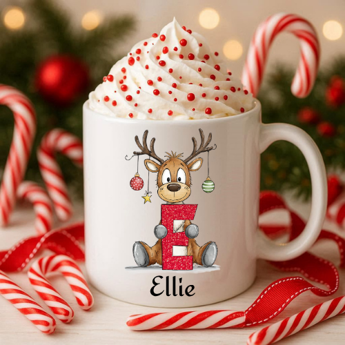 A white ceramic mug filled with whipped cream and red sprinkles, decorated with a cartoon reindeer holding a red glittery letter 'E' and the name 'Ellie' printed below. The mug is styled with candy canes, red ribbons, and holiday lights in the background.