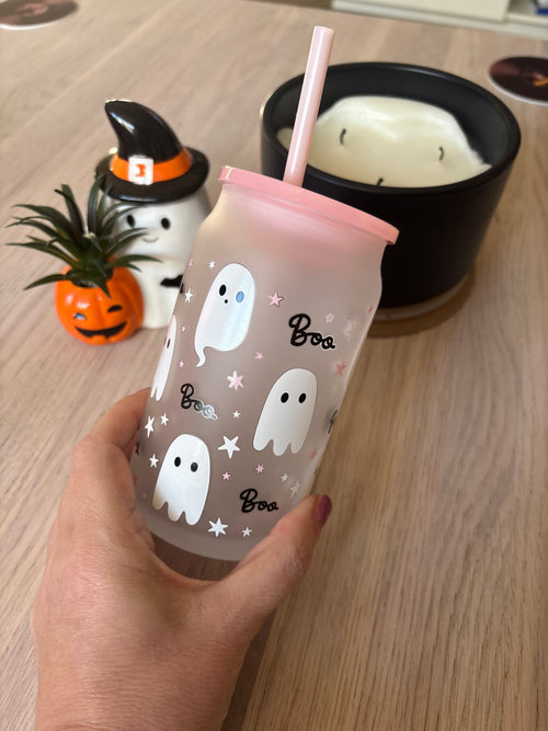 A hand holds a frosted tumbler with a pink lid and straw, decorated with white ghosts, black script text, and small pink stars. The background shows a wooden table with a small pumpkin plant, a white and black witch-themed figurine, and a black candle holder.