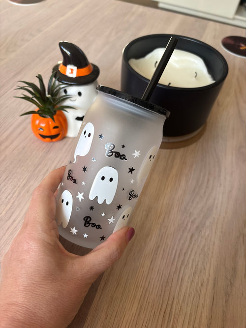 A hand holds a frosted tumbler with a black lid and straw, decorated with white ghosts, black stars, and the word 'Baa' in script. The background shows a wooden table with a black candle and a small pumpkin plant.