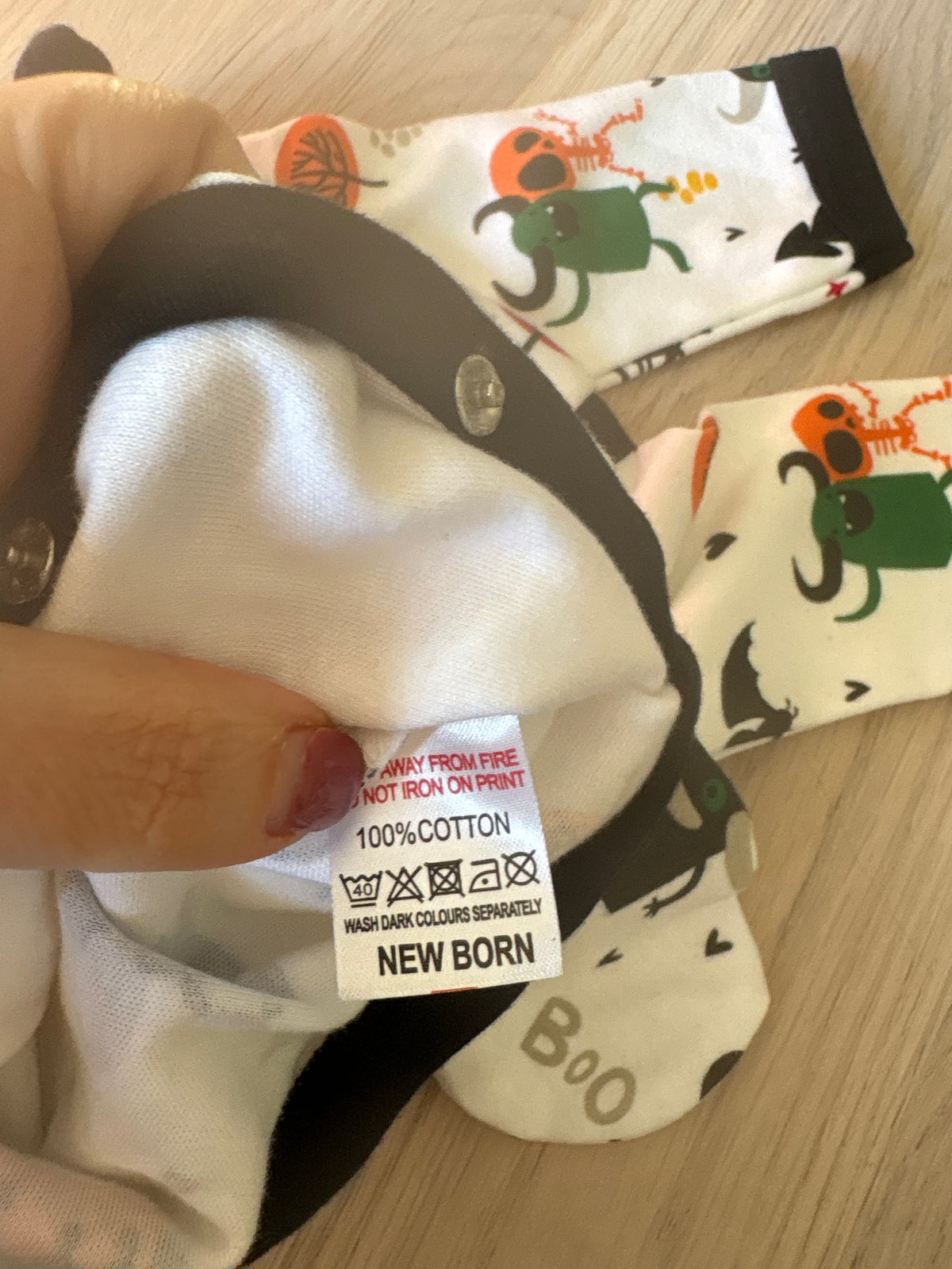 A close-up of a baby sock with a cartoon monster print, held by a hand to show the inner care label. The sock is white with a black cuff and features a green and orange monster design.