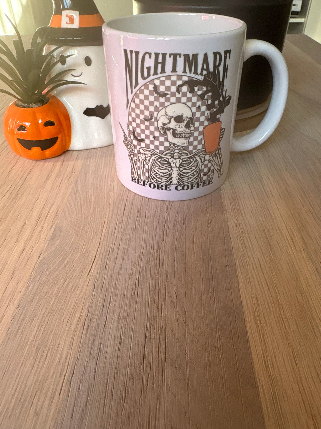 A white ceramic mug with a Halloween-themed graphic reading 'NIGHTMARE BEFORE COFFEE' featuring a skeleton holding an orange pumpkin, placed on a wooden tabletop next to a small potted plant.