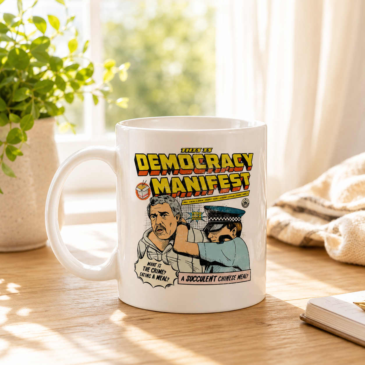 A white ceramic mug with a colorful political cartoon graphic on the side, sitting on a wooden table in a bright, sunlit room with a plant and a book nearby.