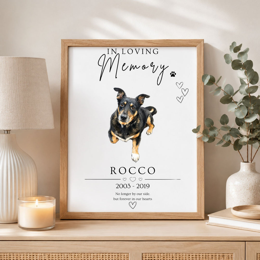 A framed memorial print featuring a watercolor-style dog portrait with the text 'In Loving Memory Rocco 2005-2019' and a short remembrance message, displayed on a tabletop in a styled room setting with a lamp, candle, and vase.