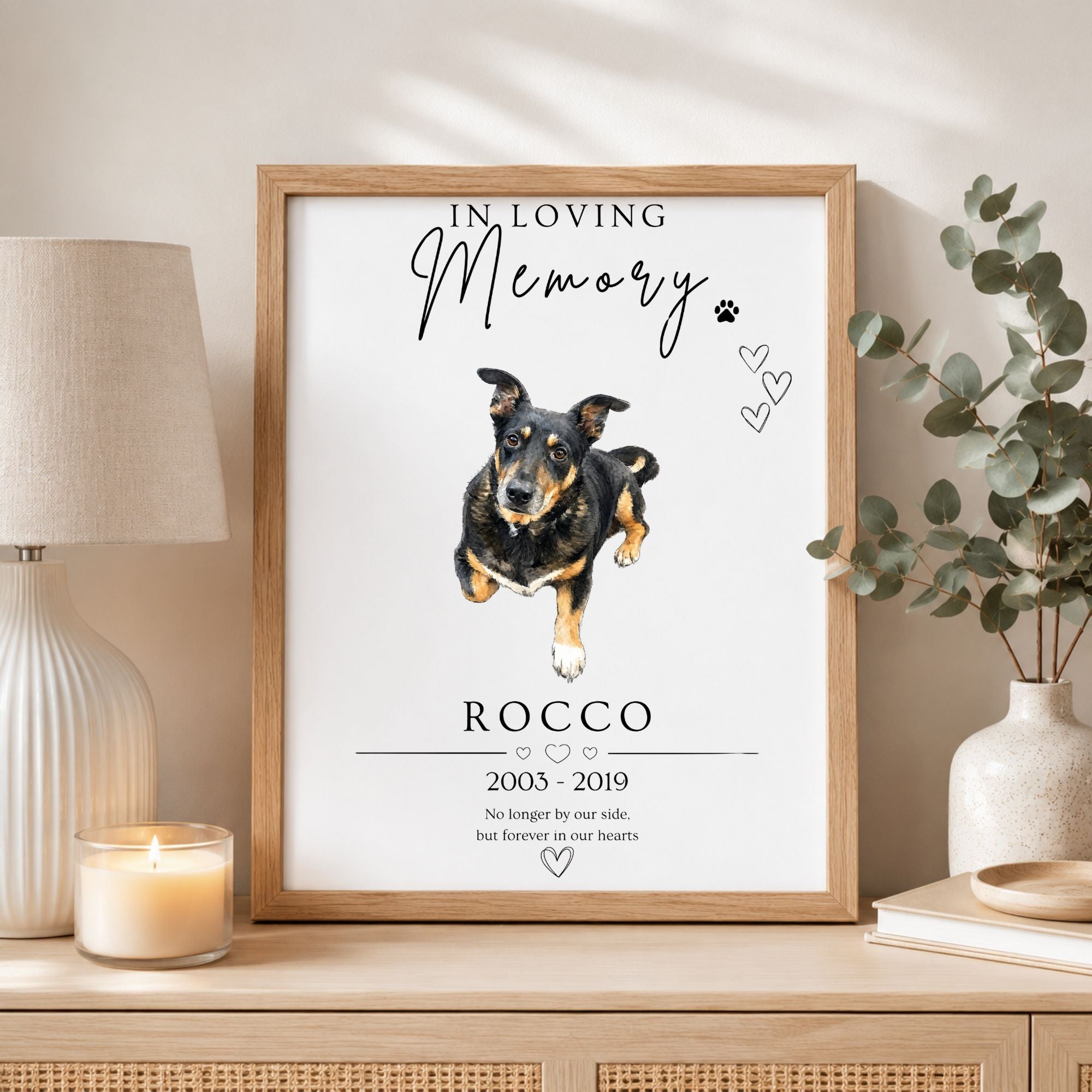 A framed memorial print featuring a watercolor-style dog portrait with the text 'In Loving Memory Rocco 2005-2019' and a short remembrance message, displayed on a tabletop in a styled room setting with a lamp, candle, and vase.