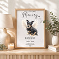 A personalized pet memorial print in a light wood frame, displayed on a wooden console table in a styled room setting with a lamp, candle, and potted eucalyptus plant. The print features a portrait of a black and tan dog with white angel wings and the text 'IN LOVING Memory Rocco 2005 - 2019'.