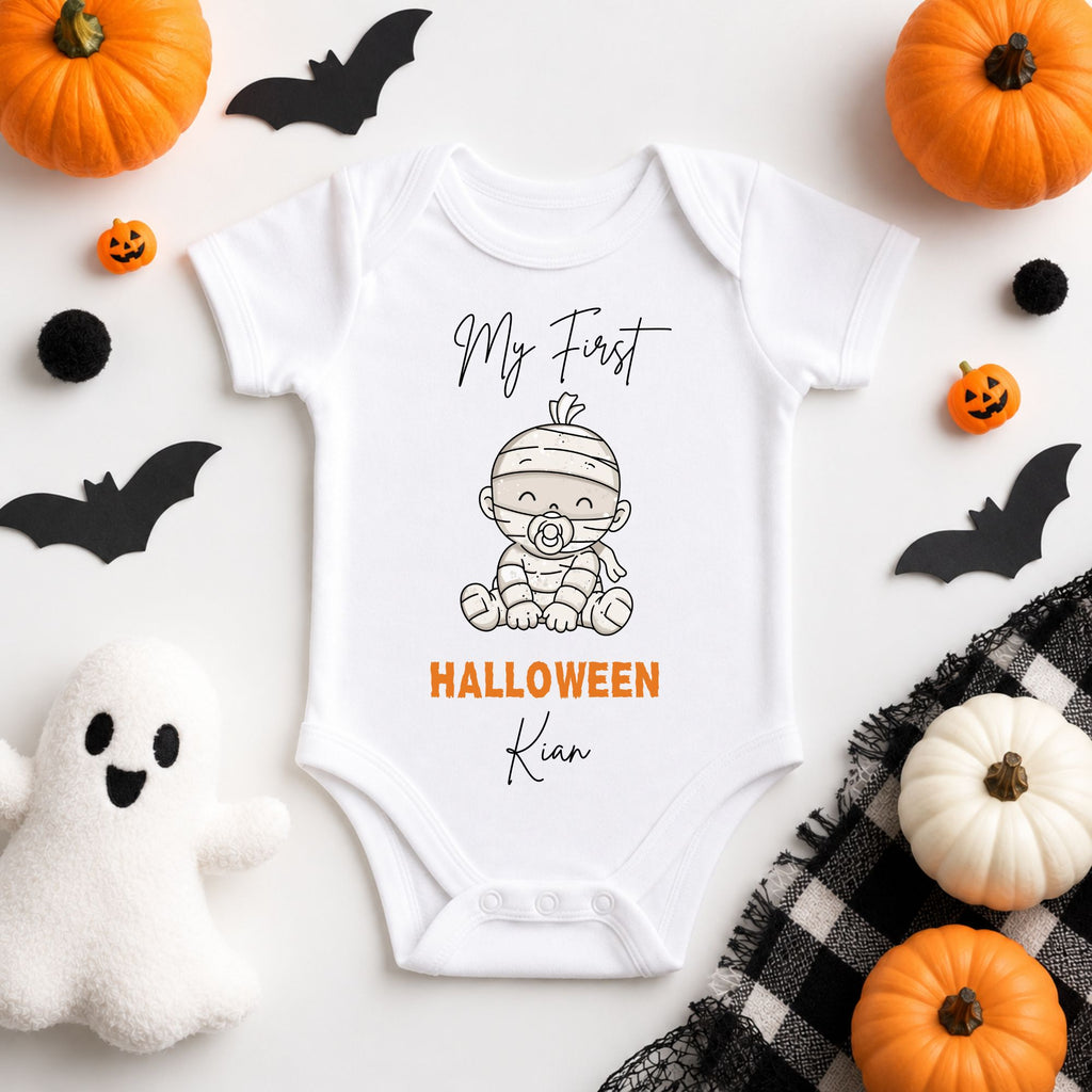A white short-sleeve baby bodysuit laid flat on a white surface, printed with a mummy graphic and the words 'My First HALLOWEEN Kia' in black and orange. The bodysuit is styled with Halloween props including pumpkins, black bat cutouts, a ghost, and a plaid cloth.