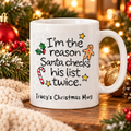 A white ceramic mug with a Christmas-themed printed message reading 'I'm the reason Santa checks his list twice.' and 'Tracy's Christmas Mug,' styled on a wooden surface with holiday greenery, ornaments, and warm blurred lights in the background.