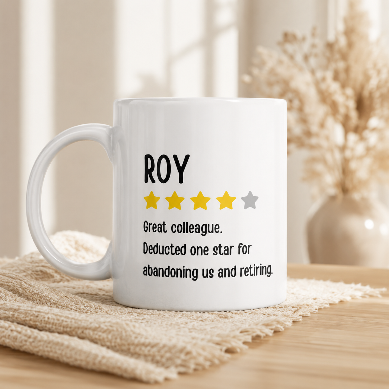 A white ceramic mug with a humorous dictionary-style definition of 'ROY' featuring yellow and gray stars, resting on a beige woven placemat on a wooden table with a blurred background of dried flowers.