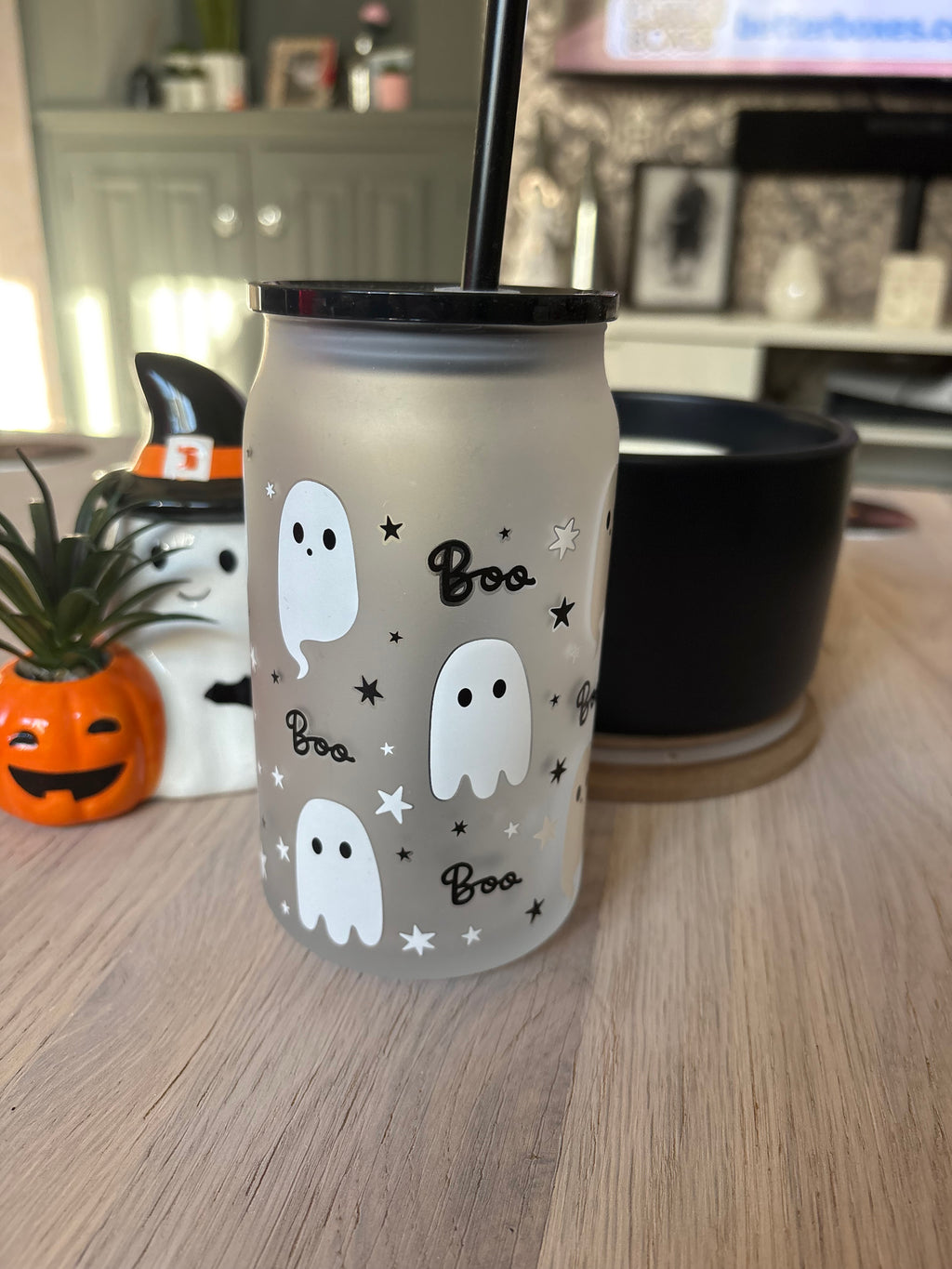 A frosted glass tumbler with a black lid and straw, decorated with white ghosts, black stars, and the word 'Boo' in script, sitting on a wooden table in a home setting.