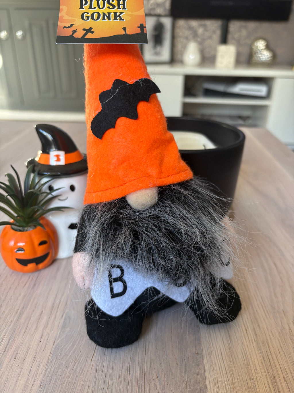 A plush Halloween gnome figurine with an orange pointed hat, black bat detail, gray beard, and white shirt with a black letter 'B', standing on a wooden table in a home setting.