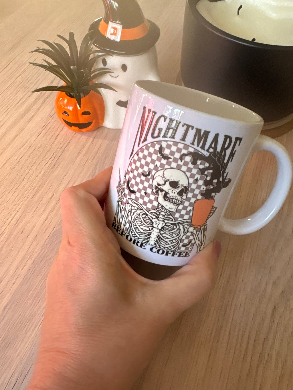 White ceramic Halloween coffee mug with a skeleton holding an orange coffee cup, bats and “Nightmare before coffee” text