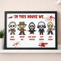 A framed horror-themed wall print hanging on a beige wall above a shelf. The print features five cartoon characters in masks with labels reading 'LOVE FAMILY', 'DREAM BIG', 'LIKE SCARY MOVIES', 'PLAY GAMES', and 'LOVE FRIDAY' beneath them, with red blood-splatter graphics and the text 'IN THIS HOUSE WE.