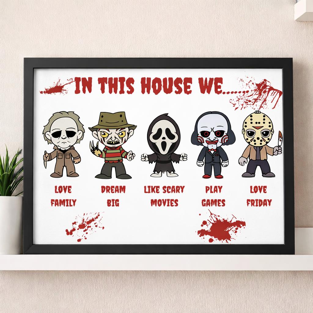 A framed horror-themed wall print hanging on a beige wall above a shelf. The print features five cartoon characters in masks with labels reading 'LOVE FAMILY', 'DREAM BIG', 'LIKE SCARY MOVIES', 'PLAY GAMES', and 'LOVE FRIDAY' beneath them, with red blood-splatter graphics and the text 'IN THIS HOUSE WE.