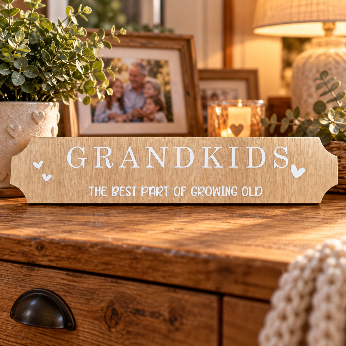 A personalized wooden sign reading 'GRANDKIDS THE BEST PART OF GROWING OLD' with small heart accents, displayed on a wooden dresser in a warmly lit home setting with framed photos and plants in the background.