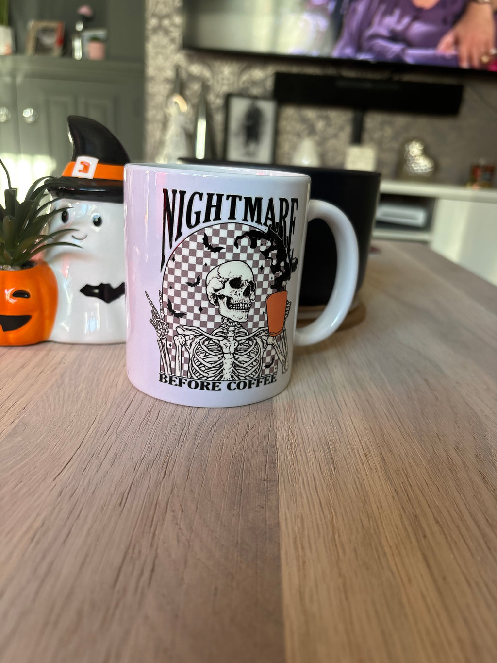 A white ceramic mug with a black and orange 'Nightmare Before Coffee' skeleton graphic sits on a wooden table in a living room setting.