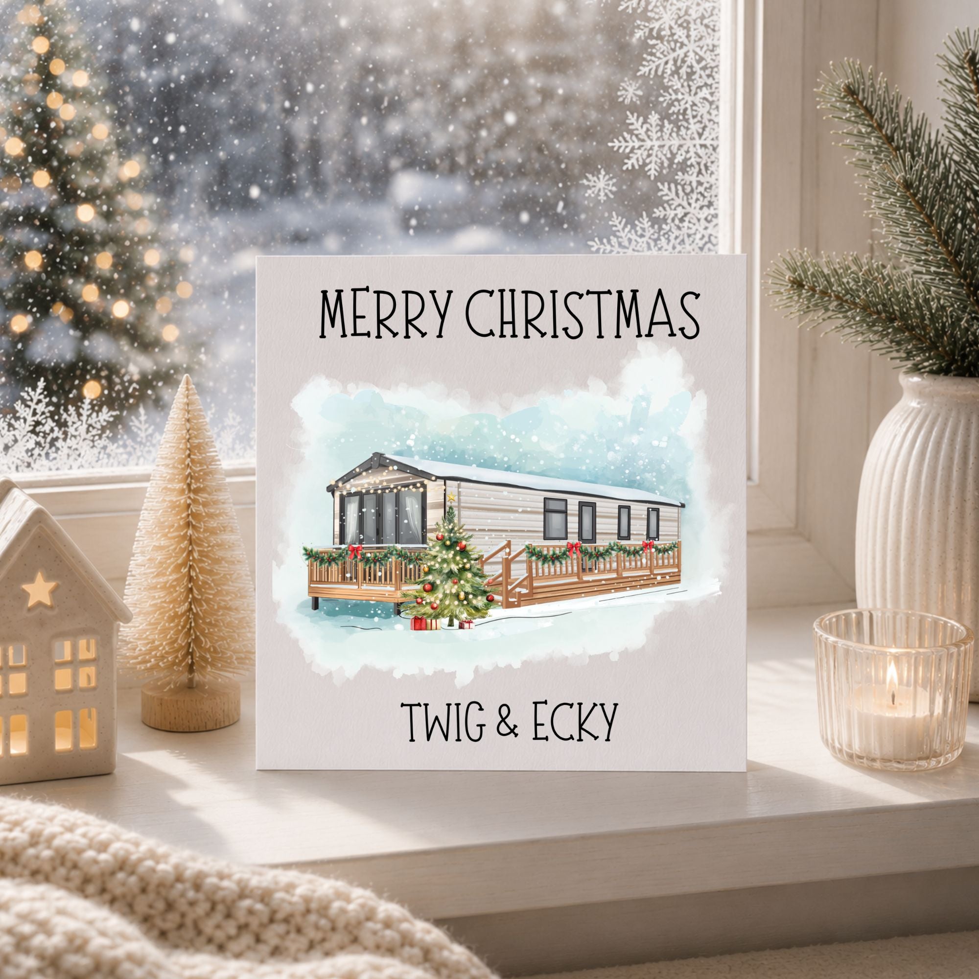 A square Christmas greeting card featuring a watercolor illustration of a caravan with a decorated Christmas tree on a snowy porch, with the text 'MERRY CHRISTMAS' at the top and 'TWIG & ECKY' at the bottom. The card is styled on a windowsill with holiday decorations, including a small lit house, a miniature tree, a candle, and a snowy window view in the background.